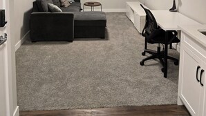 Ein kompakter Raum mit einem grauen Ecksofa, einem kleinen runden Tisch, einem Schreibtisch mit Stuhl, einem an der Wand befestigten Fernseher und einem weißen Schrank.