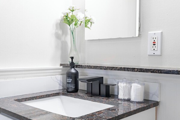 A bathroom with a white sink, a black soap dispenser, a vase with white flowers, and a power outlet on the wall.