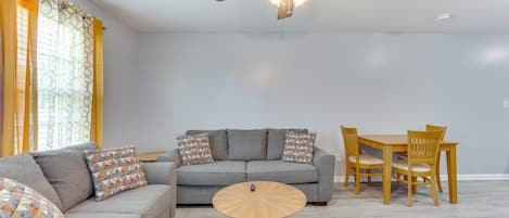 A living room with a grey sofa, patterned cushions, a wooden coffee table, and a dining area with wooden chairs and a table.