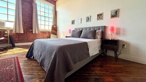 A bedroom with a large bed, wooden flooring, a brick wall, and a window with curtains. - Downtown: The Belle Isle View Loft (Detroit)