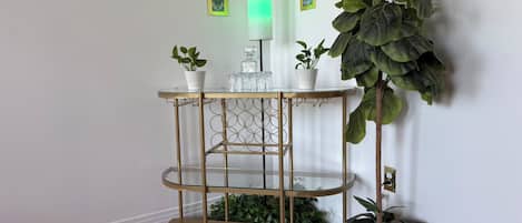 A corner with a gold metal shelf unit holding decorative items, a green wall lamp, and a large potted plant.