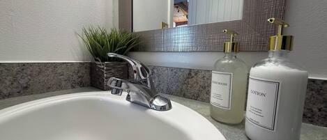 Un baño con un lavamanos blanco, un grifo moderno, un espejo y dos botellas de jabón de manos y loción.