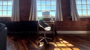 A room with a wooden floor, a desk with a computer, and a chair. There are two windows with curtains, and a brick wall. - Downtown: The Belle Isle View Loft (Detroit)