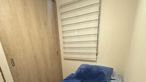 Ein kleines Zimmer mit einem Einzelbett, einer Holztür, einem Fenster mit Jalousien und einer blauen Bettdecke.