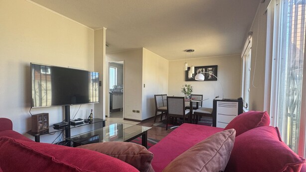 Ein Wohnzimmer mit Flachbildfernseher, einem Glastisch und roten Sofas. Es gibt eine Essecke mit Tisch und Stühlen, und im Hintergrund eine Küche.