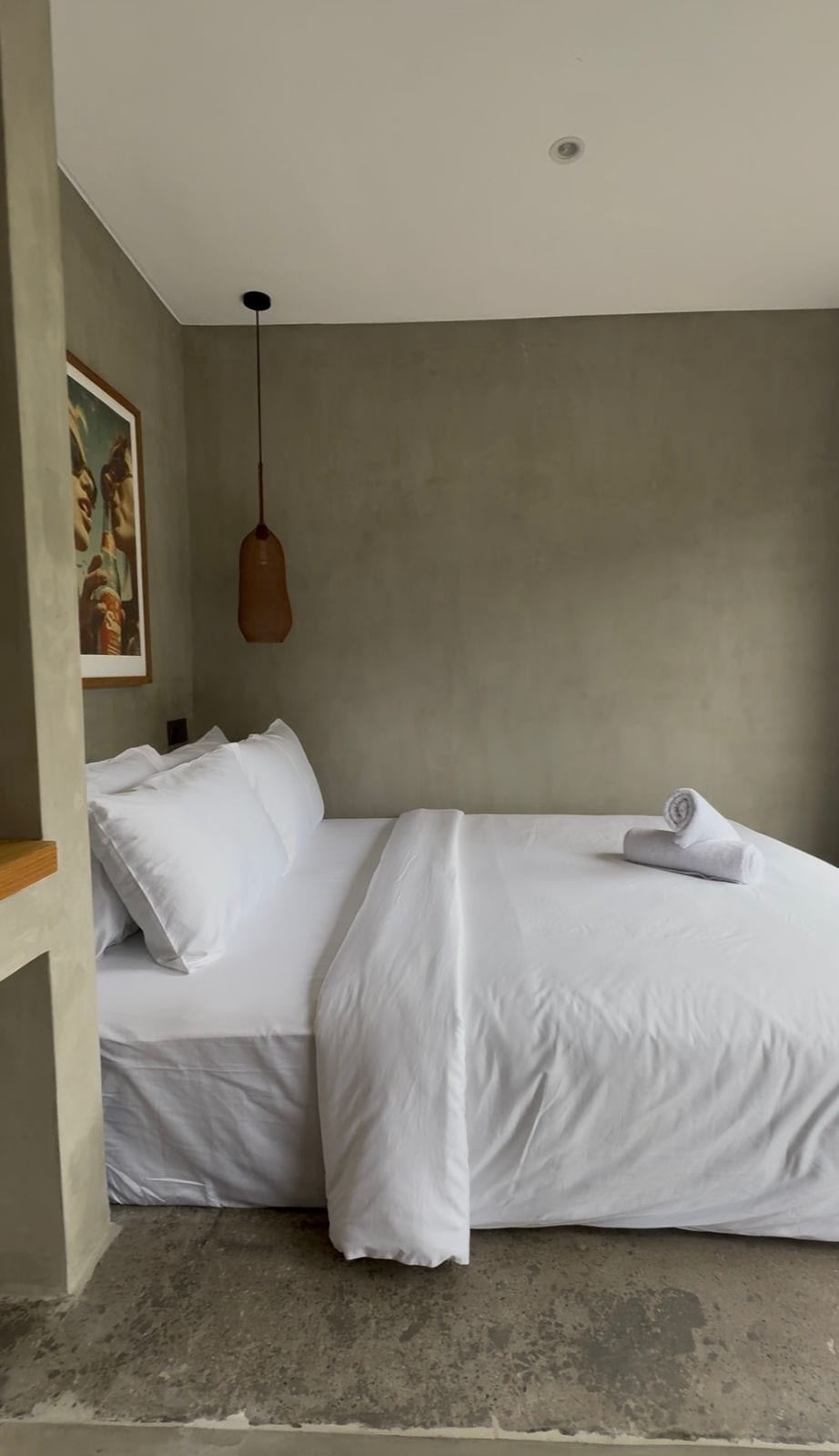 A bedroom with a bed, pillows, a rolled towel, a pendant light, and a framed picture on the wall.