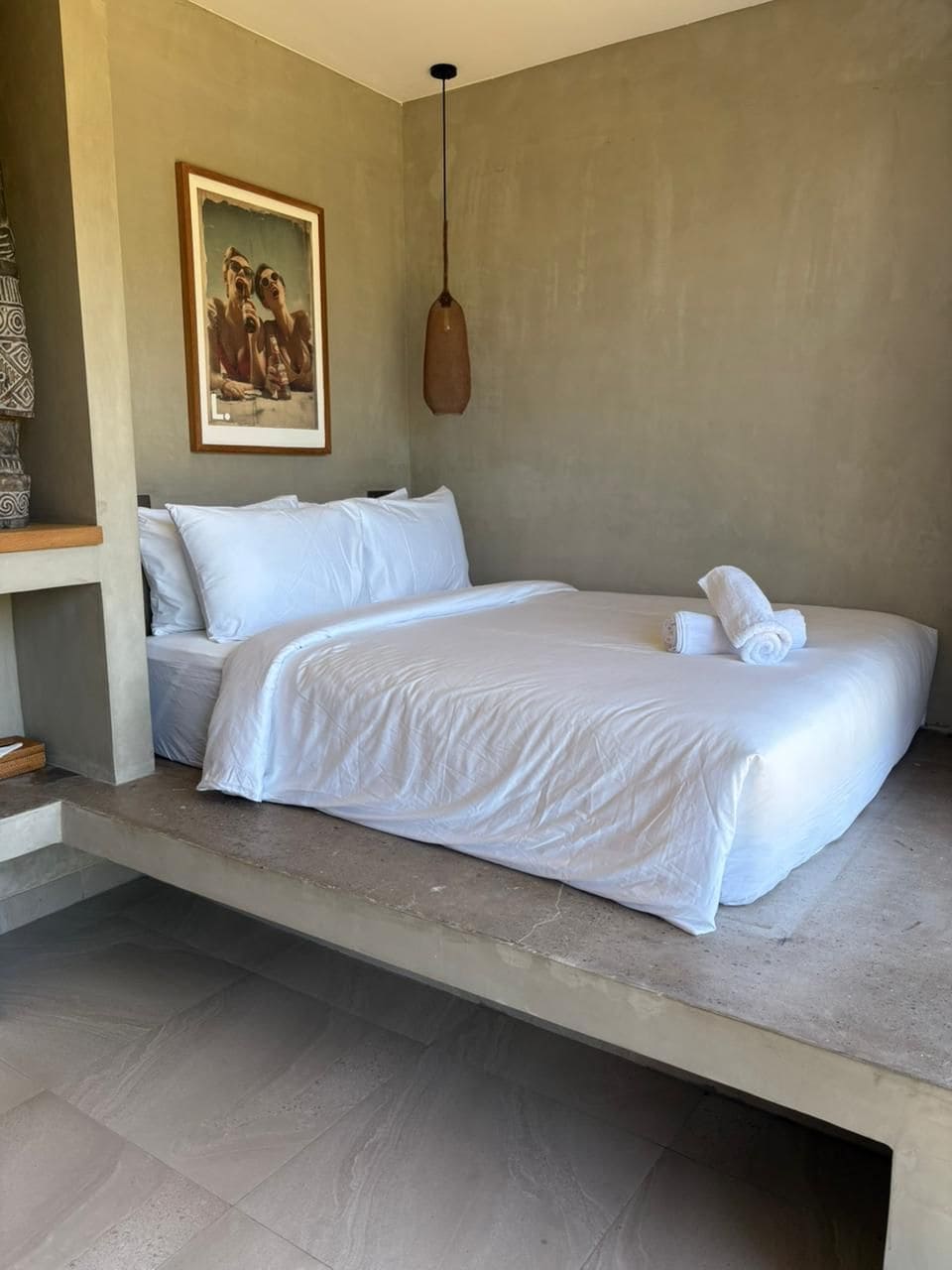 A hotel room with a bed, a painting on the wall, and a hanging light fixture.