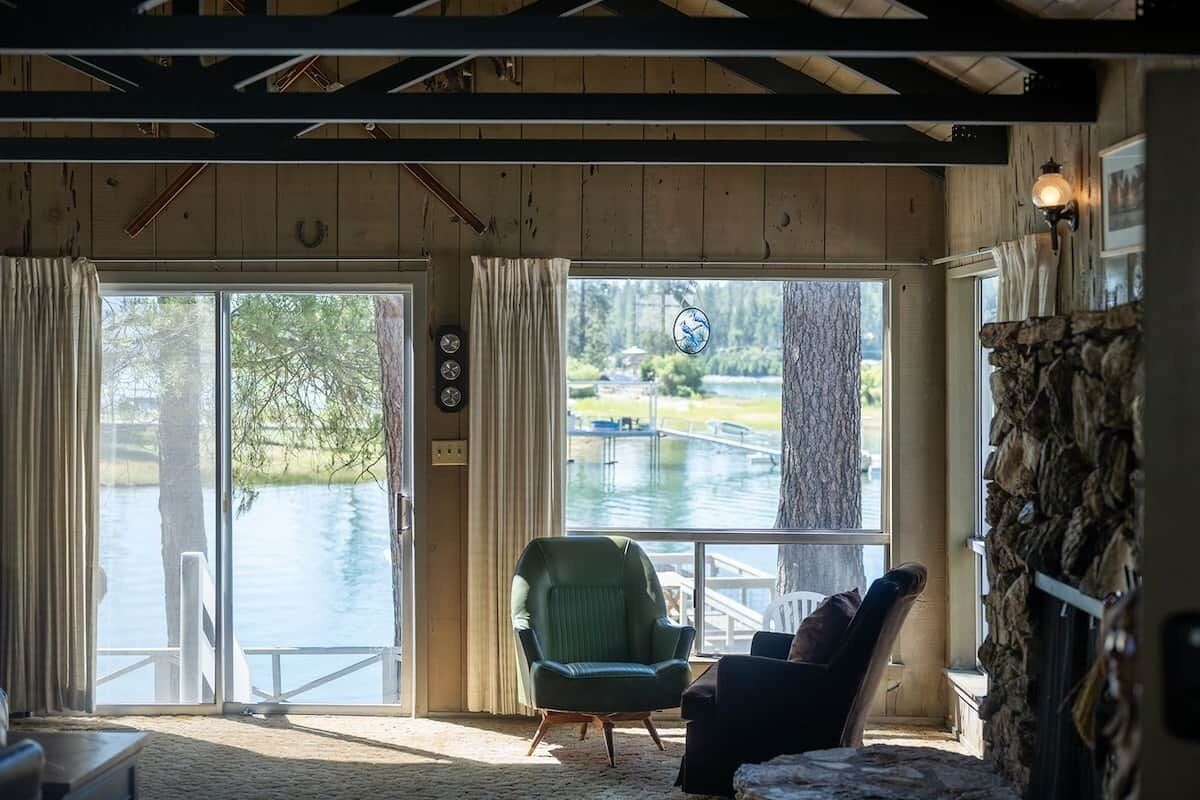 Huoneessa on suuri ikkuna, josta avautuu näkymä vesistöön. Siellä on myös vihreä nojatuoli, musta nojatuoli ja kiviseinä.