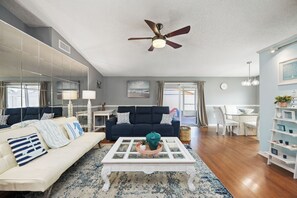 A spacious living room with a white sofa, a glass coffee table, a dark blue couch, and a ceiling fan.