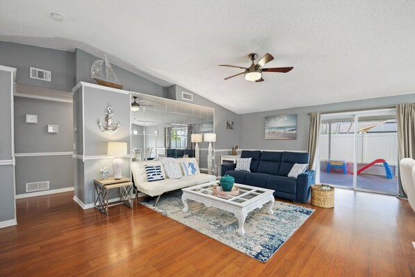 A spacious living room with a white sofa, a blue couch, and a coffee table. There is a ceiling fan and a large window with curtains.