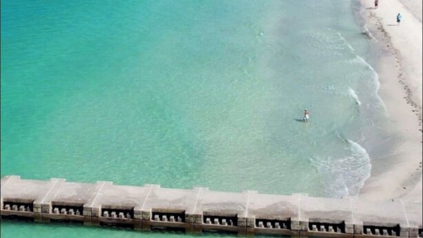 A beach with clear turquoise water, a sandy shore, and a few people. There are breakwaters in the water.