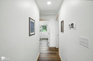 A hallway with white walls, wooden flooring, and framed artwork. There are light switches and a thermostat on the wall.