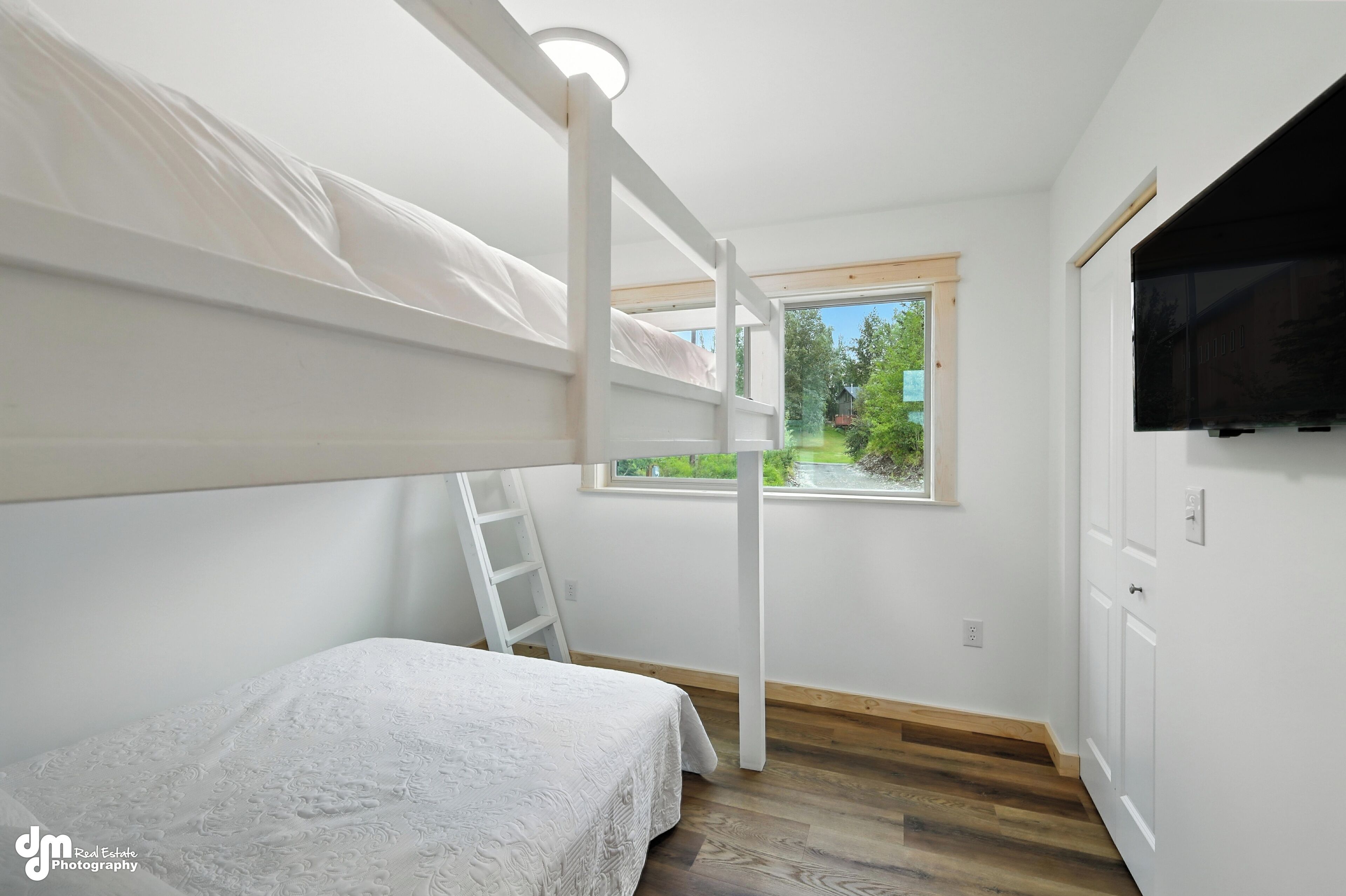A bedroom with a bunk bed, a window with a view of greenery, a TV mounted on the wall, and a wooden floor.