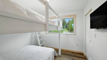 A bedroom with a bunk bed, a window with a view of greenery, a TV mounted on the wall, and a wooden floor.
