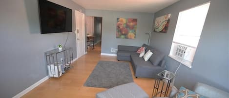 A living room with a grey sofa, a grey ottoman, a flat-screen TV mounted on the wall, and a painting on the wall.