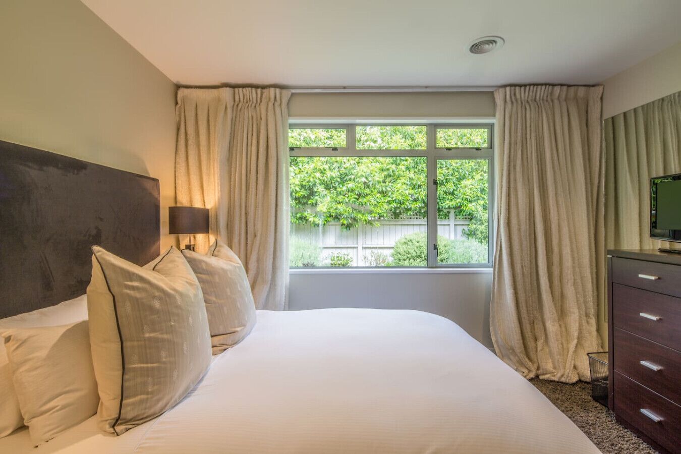 Een slaapkamer met een bed, nachtkastje, lamp, dressoir en een raam met gordijnen.