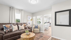 A modern living room with a brown leather sofa, a round wooden coffee table, and a beige armchair. There is a large framed picture on the wall and a door with a black frame. - Cozy 3BR Getaway with Hot Tub, Firepit & BBQ (Clinton Township)