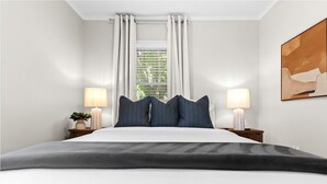 A modern bedroom with a large bed, two bedside lamps, a window with curtains, and a painting on the wall. - Cozy 3BR Getaway with Hot Tub, Firepit & BBQ (Clinton Township)