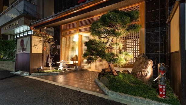 Un edifici tradicional japonès amb entrada de fusta, un petit jardí amb un bonsai i un fanal de pedra.