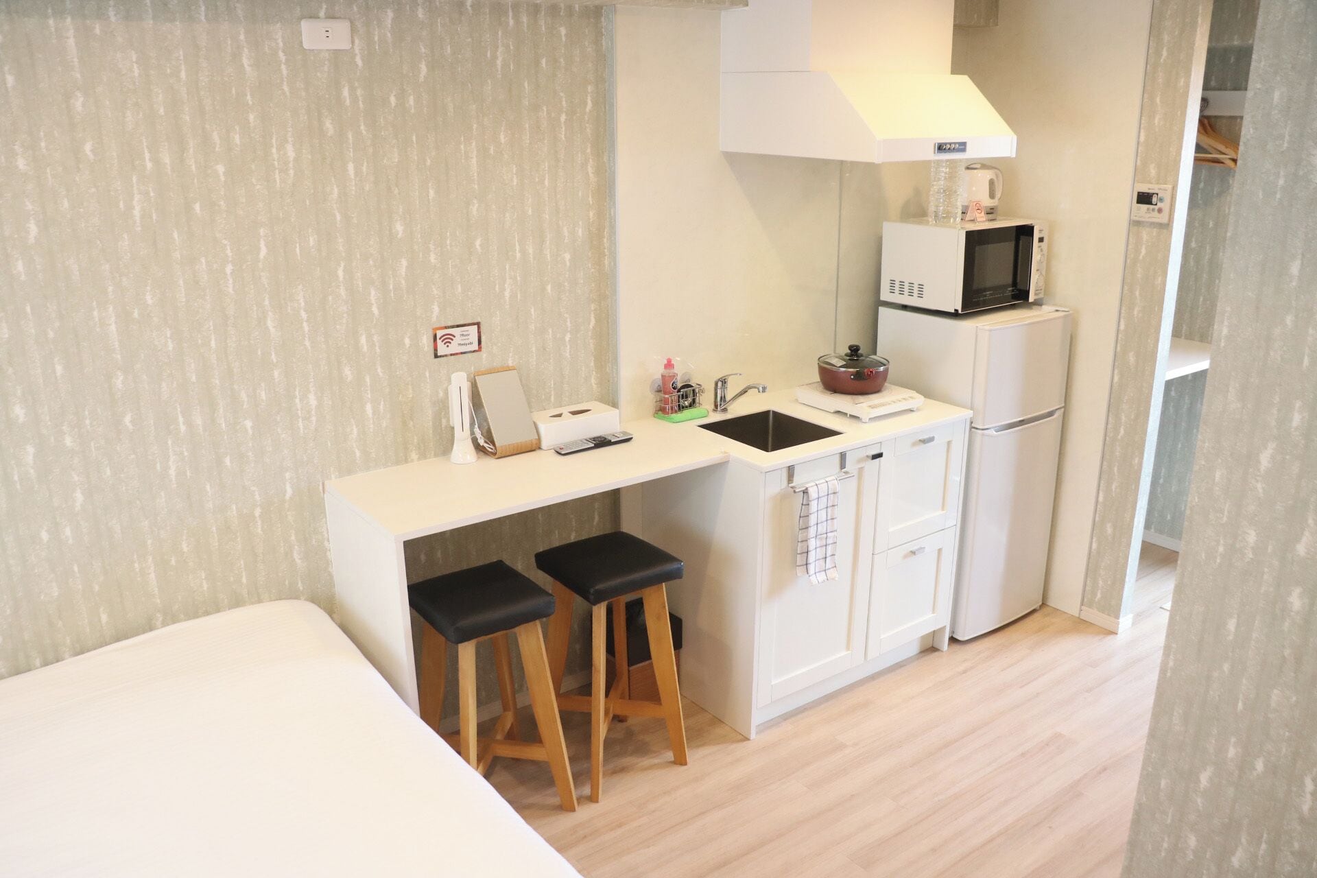 A compact kitchenette with a microwave, sink, and small refrigerator. There are two stools and a small table with a kettle and a kettle pot.
