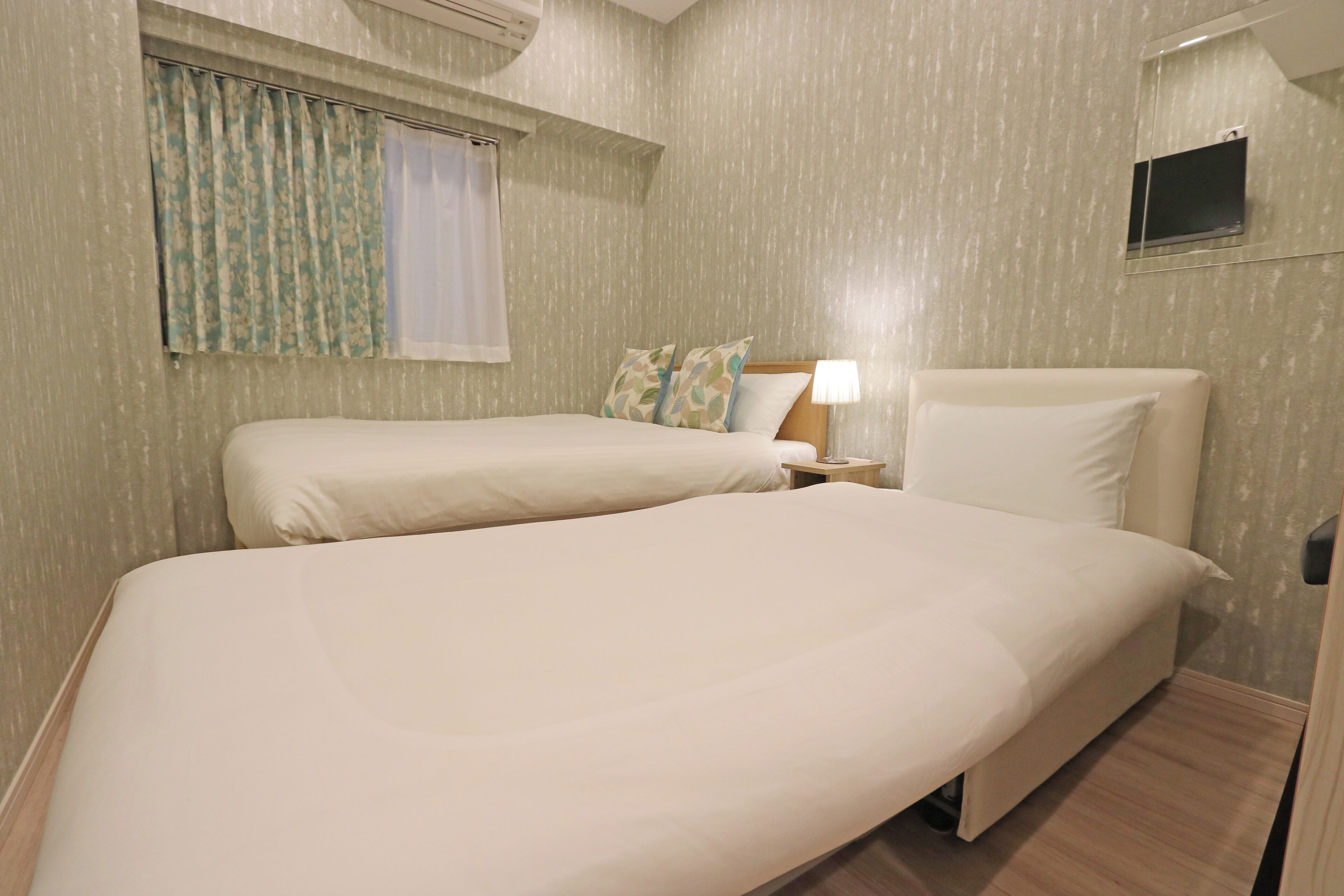 A hotel room with two beds, a small bedside table with a lamp, a window with curtains, and a wall-mounted TV.