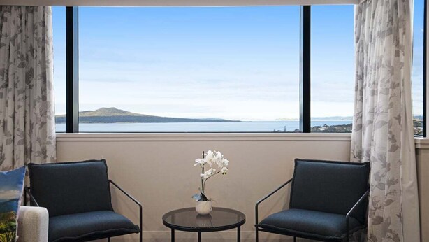 Ein Zimmer mit zwei Stühlen, einem kleinen Tisch und einer Vase mit Blumen. Ein großes Fenster bietet Blick auf ein Gewässer und eine entfernte Landschaft.