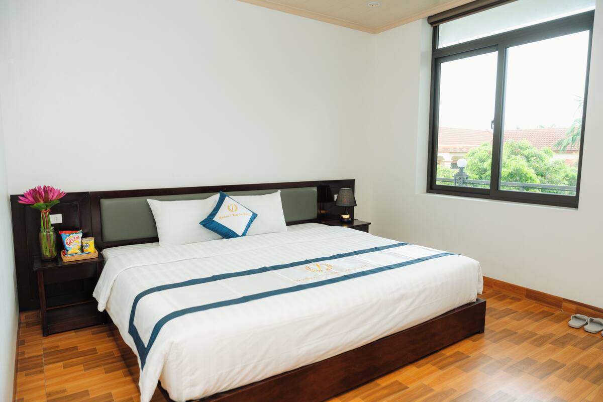 A neatly made bed with a white and blue striped bedspread, a nightstand with a lamp, and a window with a view of greenery.