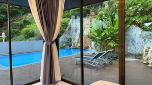 Une chambre dotée d’une grande porte-fenêtre coulissante donnant sur une piscine, avec un lit et une vue sur un jardin aménagé, agrémenté d’une cascade et de statues.