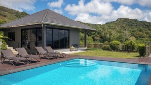 Une maison moderne dotée d’une piscine, de chaises longues et offrant une vue sur une colline boisée.