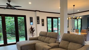 Un salon spacieux avec un canapé d’angle, une table basse en verre et un ventilateur de plafond. De grandes fenêtres offrent une vue sur l’extérieur.