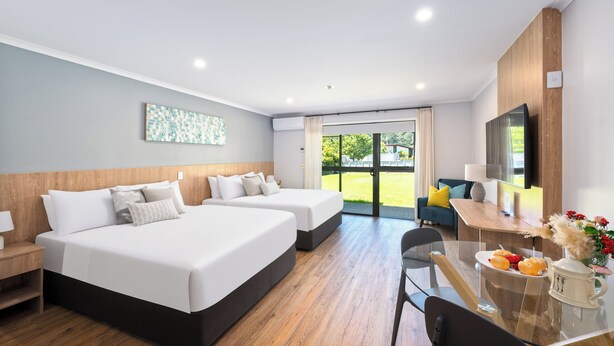 A hotel room with two beds, a desk with a chair, a TV, and a window with a view of greenery. - Albany Oak Motel (Auckland)