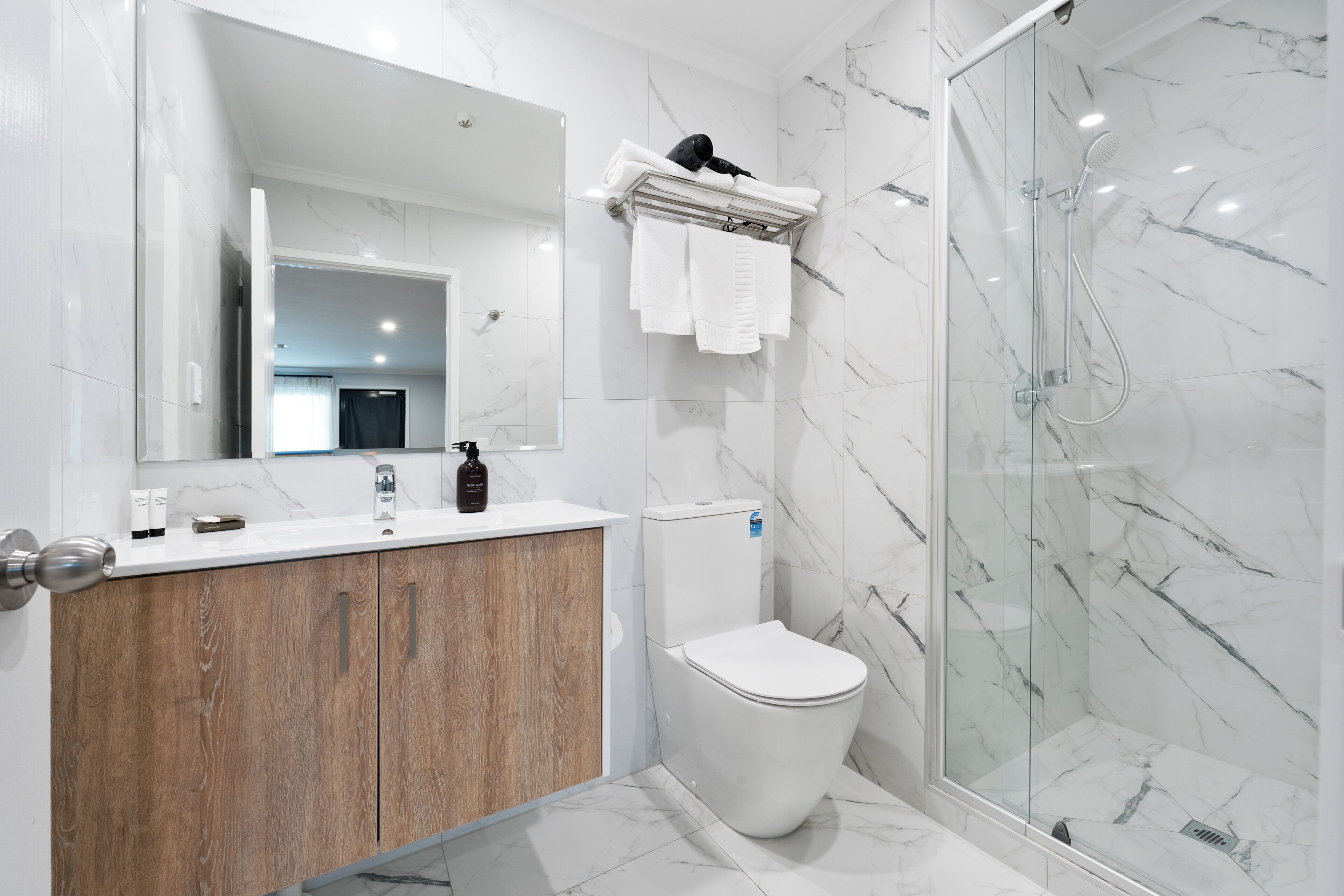 A modern bathroom with a marble shower, a wooden vanity, a toilet, and a large mirror.
