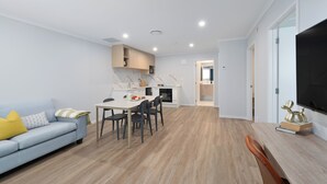 A modern living room with a sofa, dining table, and chairs. There is a kitchen area with cabinets and a microwave. The room has wooden flooring and white walls. - Albany Oak Motel (Auckland)