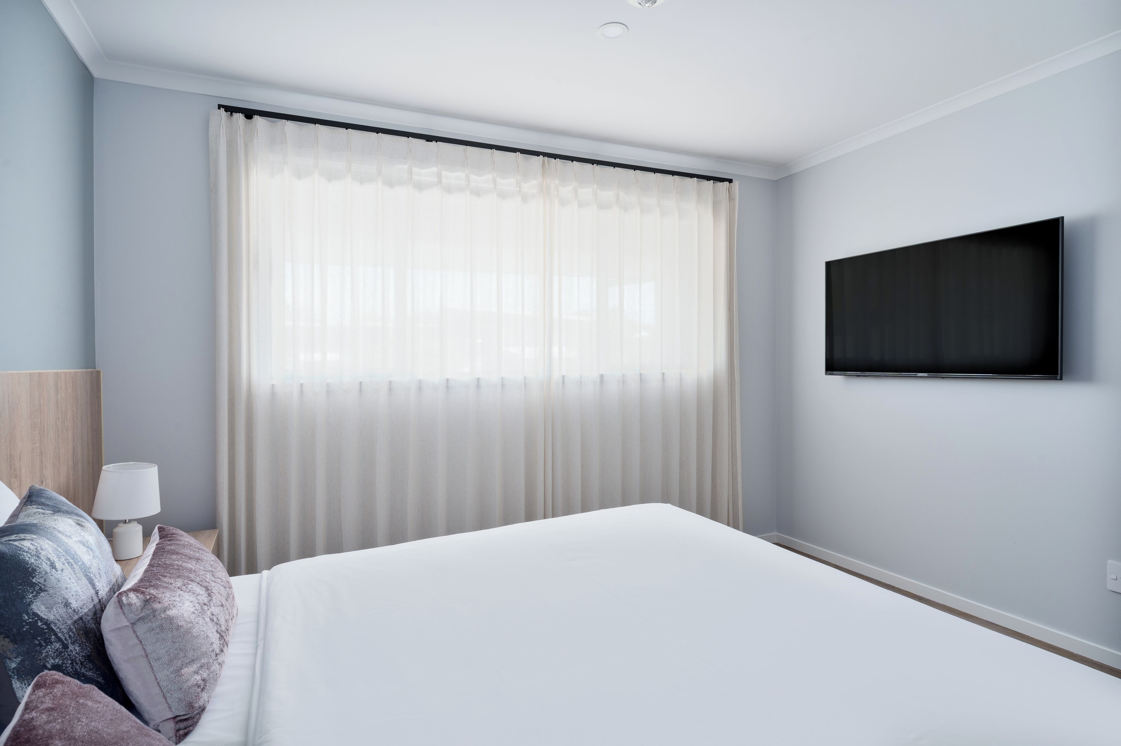 A modern hotel room with a large bed, a flat-screen TV mounted on the wall, and a window with sheer curtains.