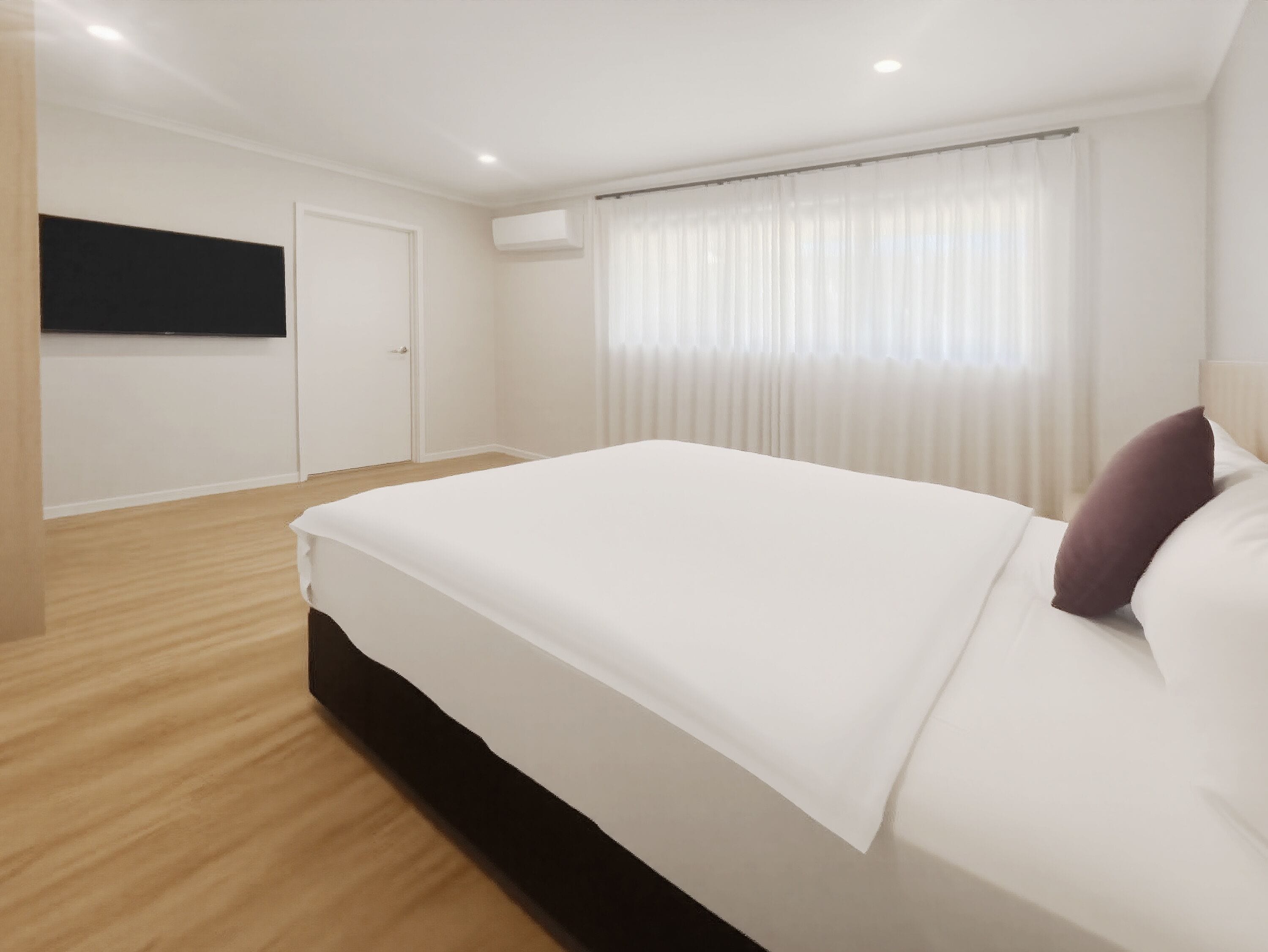 A modern hotel room with a large bed, a flat-screen TV mounted on the wall, and a window with sheer curtains.