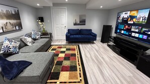A modern living room with a grey sofa, blue sofa, and a large flat-screen TV displaying various apps. - Whitby Hideout - 3BR 5BD 3WR Pool Table Sauna close to Thermea SPA (Whitby)