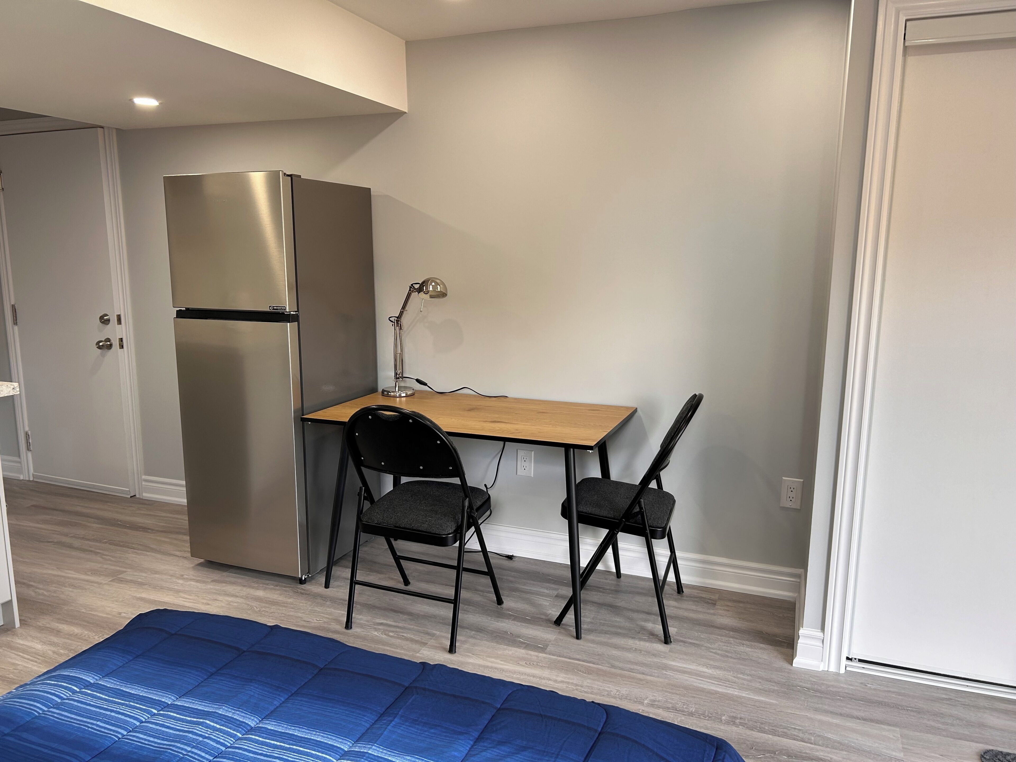 Een kleine kamer met een kitchenette, een houten bureau, twee stoelen en een blauwe mat op de vloer.