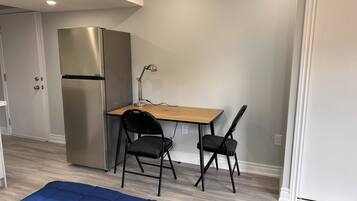 Een kleine kamer met een kitchenette, een houten bureau, twee stoelen en een blauwe mat op de vloer.