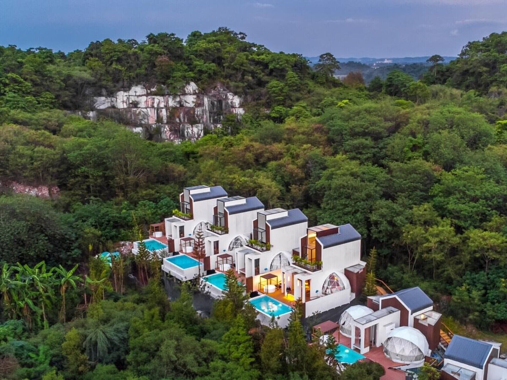 A modern, multi-story villa with a pool, surrounded by dense forest and a cliff.
