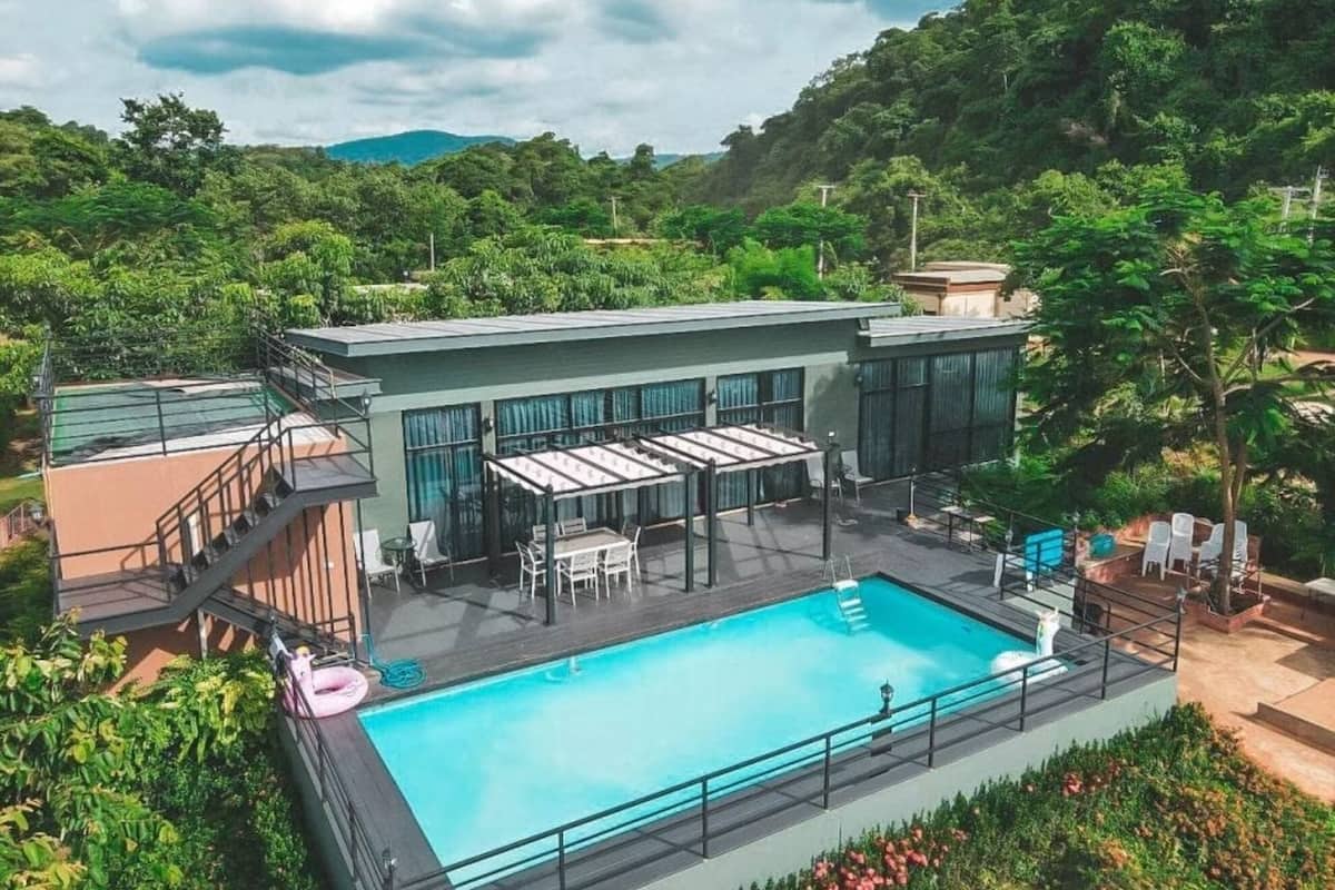 A rooftop pool area with a glass balustrade, outdoor seating, and a view of the surrounding greenery.