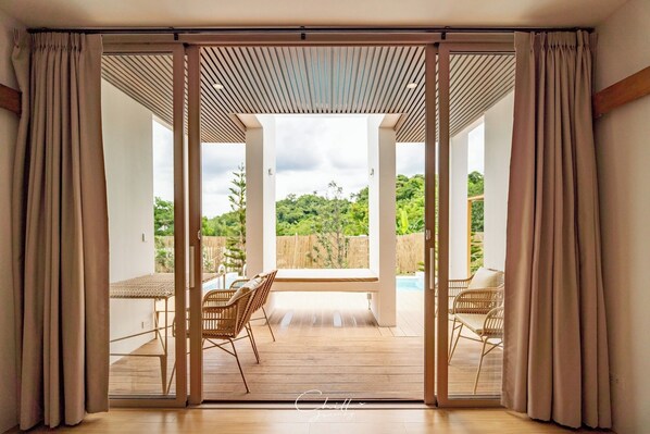 A spacious patio with a wooden table and two wicker chairs, overlooking a lush garden and a pool.