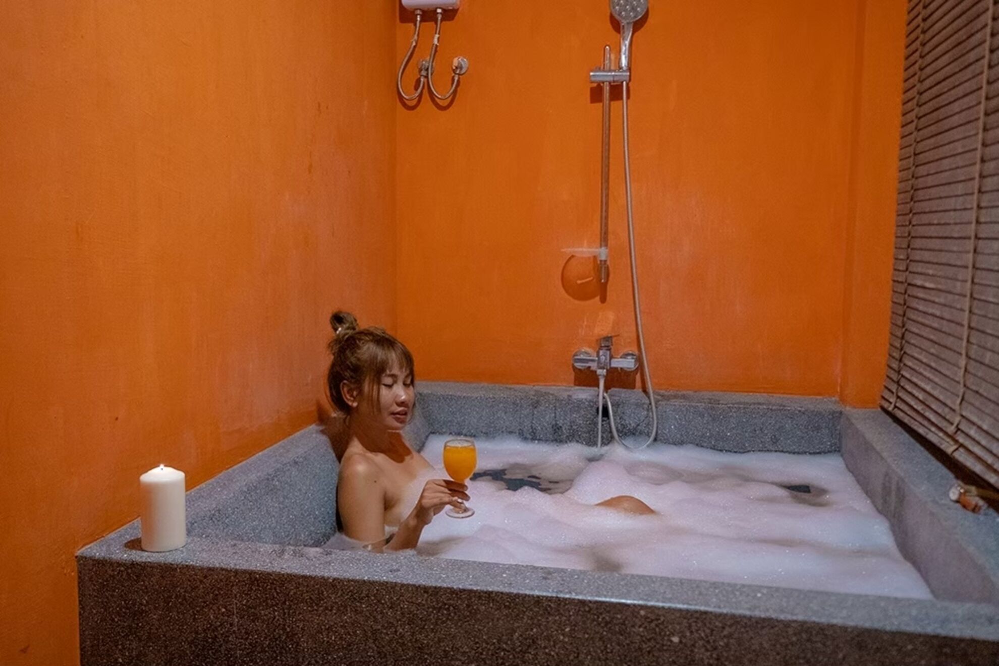 Une personne dans une baignoire, avec un verre de jus d’orange, une bougie et un pommeau de douche.