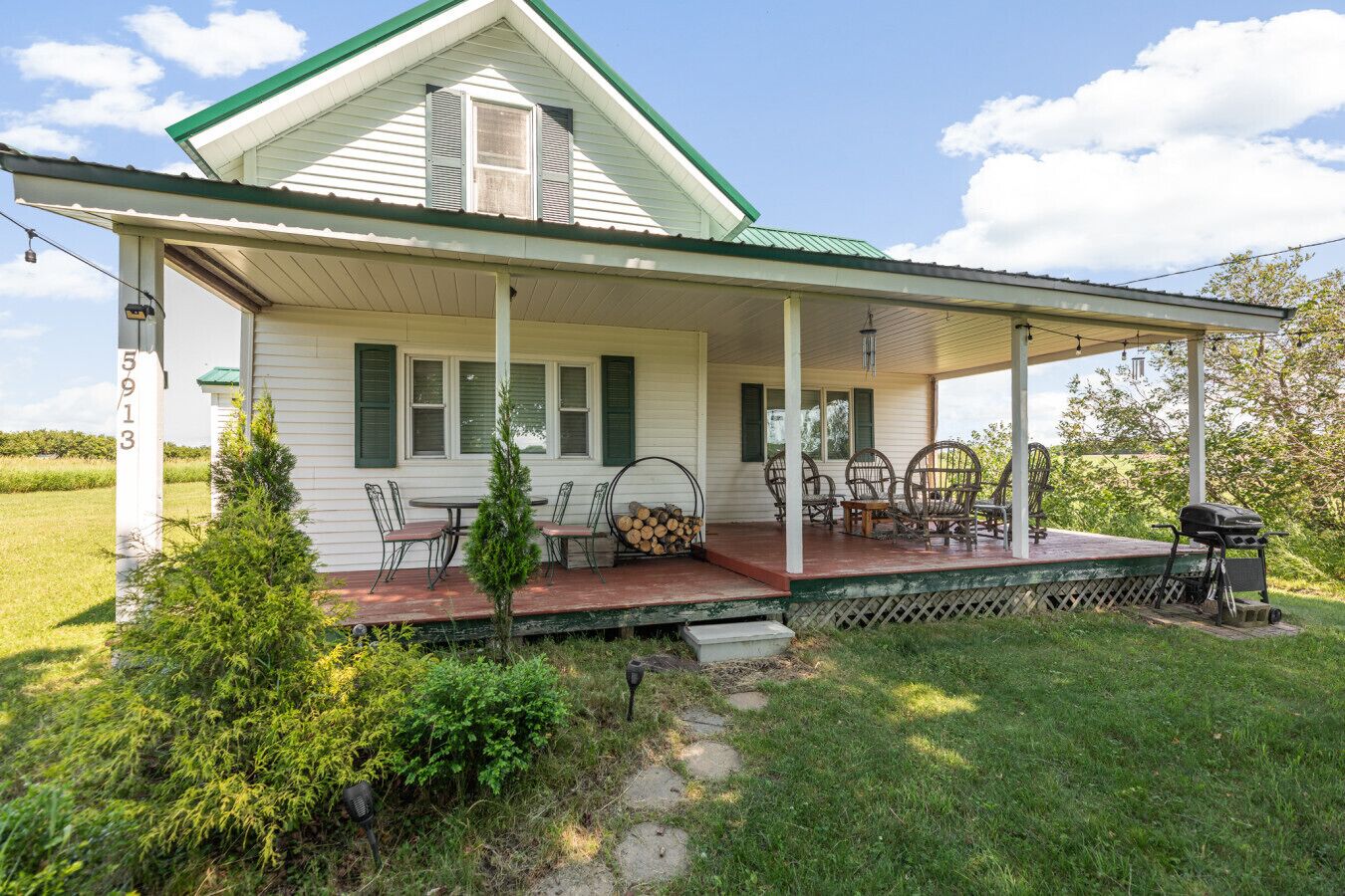 A white house with green shutters, a covered porch, and a barbecue grill.