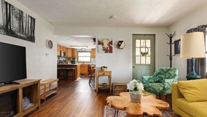 Un salon avec un téléviseur, des meubles en bois, une porte verte et une cuisine à l’arrière-plan.