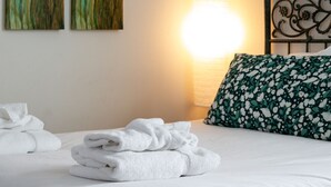 Un lit bien fait, avec du linge de lit blanc et un coussin à motifs floraux verts, une pile de serviettes blanches et deux tableaux représentant des fleurs rouges et jaunes accrochés au mur.