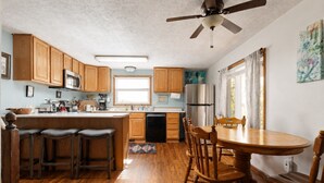 Une cuisine avec des meubles de rangement en bois, un îlot central doté de tabourets, une table à manger avec des chaises, un ventilateur de plafond et un réfrigérateur.