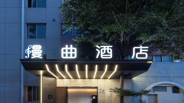 Entrada de hotel con señalización iluminada, acceso cubierto y un árbol frente a ella.