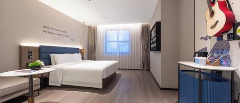 A modern hotel room with a bed, a small table, a desk, a window with curtains, and a wall with a guitar and a quote.