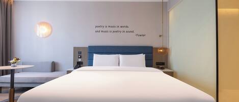 A modern hotel room with a large bed, a bedside table, a lamp, and a wall quote.