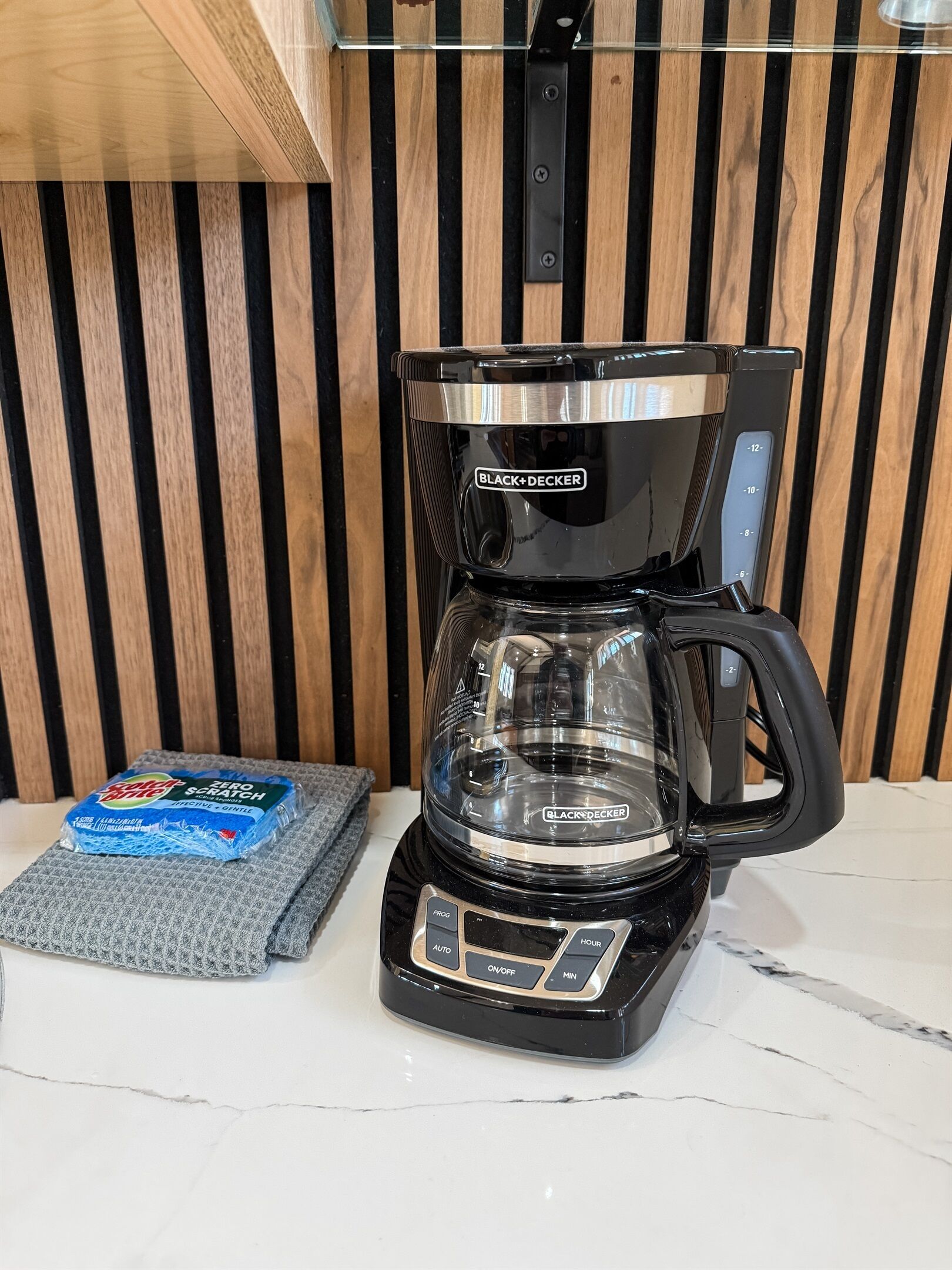 A BLACK+DECKER coffee maker with a clear carafe, a control panel with buttons, and a pack of Solo brand paper towels on a gray mat.